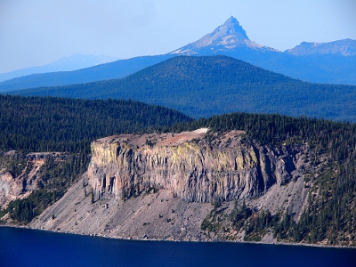 [A sliver of lake in the foreground with yellow tints on the caldera walls. Next level back is a green, tree-covered mountain. In the background is a rock summit.]